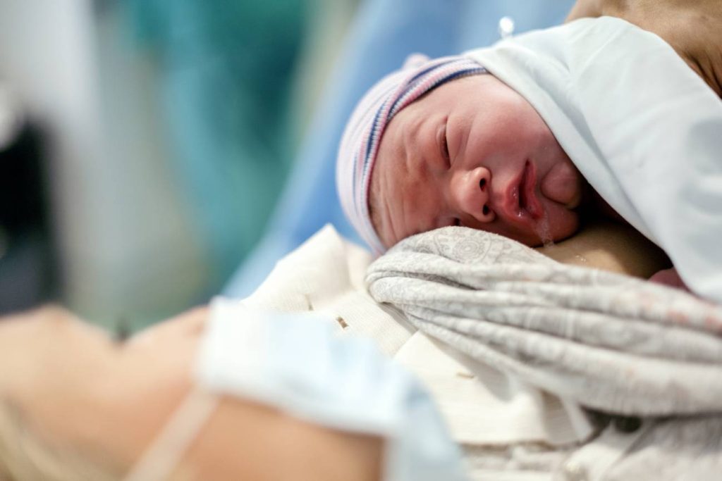 Una mujer con cubrebocas mira a su bebé recién nacido- Adobe Stock
